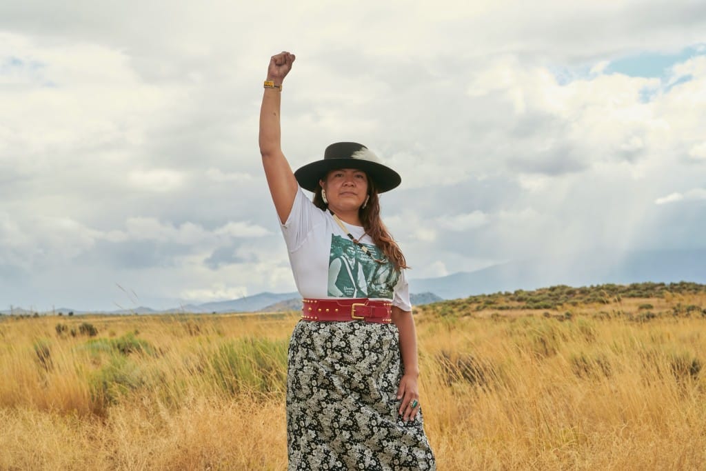 A photograph of tribal rights activist Bethany Sam, standing outside, with a raised fist